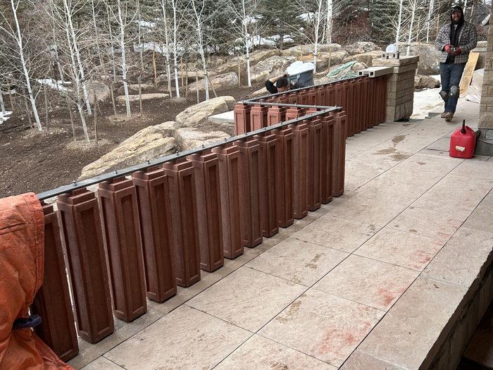 Balusters mounted to the dark metal top rail ahead of the stone caps, crew on the balcony.